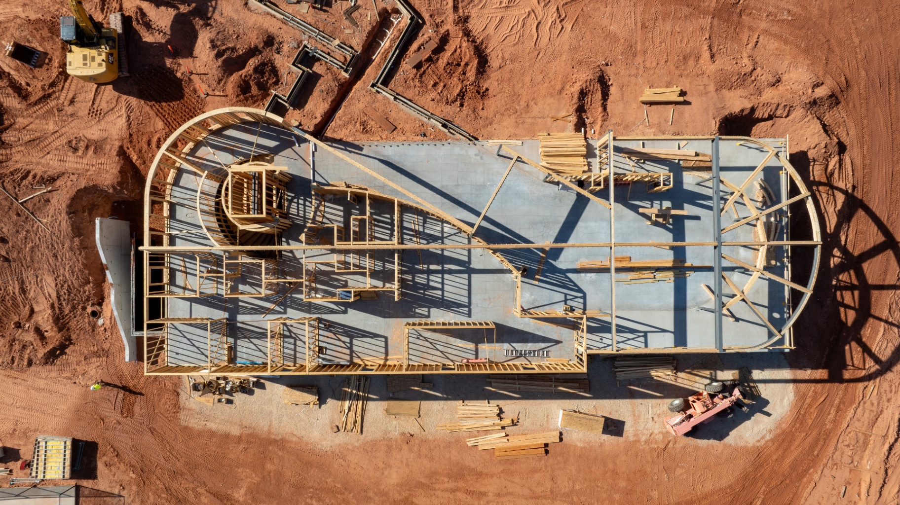 Top-down aerial drone view of a commercial Southern Utah jobsite showing framing, concrete slab, excavator, and material stockpiles around an active structure